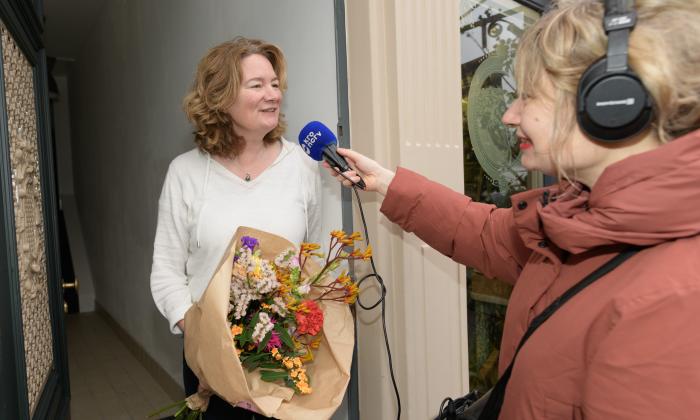 Nienke Meijer in deuropening met bos bloemen die wordt geinterviewd door verslaggever in roodbruine jas met microfoon in haar hand