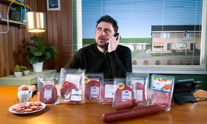 Verslaggeve Ersin Keris met telefoon in hand en voor hem liggen diverse pakjes voorverpakte cervelaat en salami-plakjes